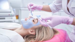 Beautician spreading blue clay paste over woman face using pair of brushes